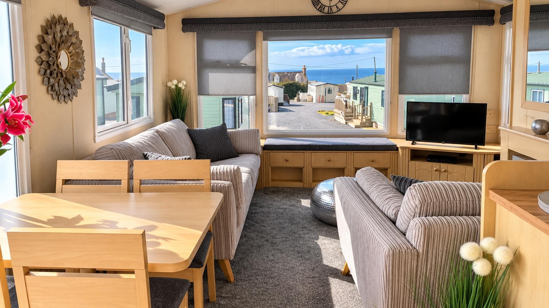 L'intérieur d'une caravane comprenant un coin repas, des sièges, un téléviseur et une vue sur un village côtier.