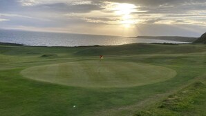 Golfkenttä, jonka keskellä on lipunvarsi, ja jota ympäröi vihreä nurmikko. Taustalla näkyy vesialue.