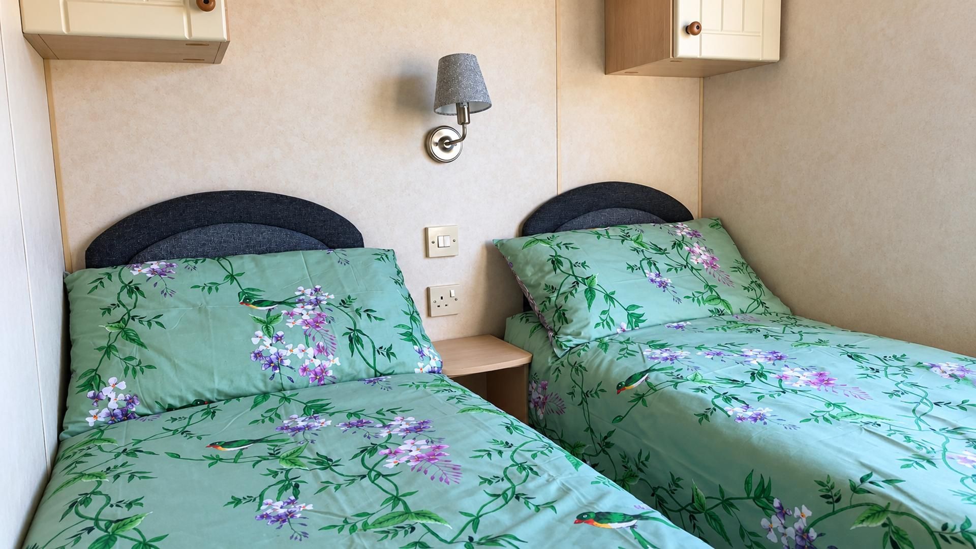 A bedroom with two beds, each with floral bedding, a bedside table, a lamp, and a wall-mounted switch.