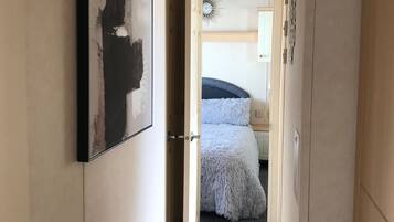 A narrow hallway leading to a bedroom with a bed, a wall-mounted clock, and a door with a handle.