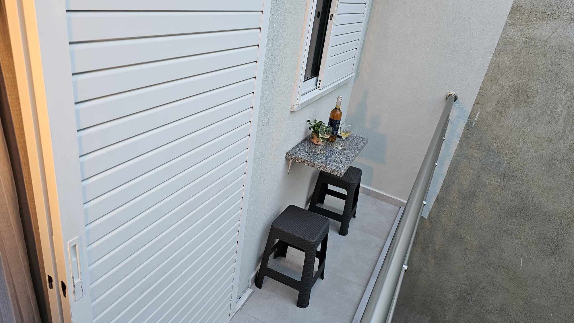 A small outdoor seating area with a table and two chairs, a window with blinds, and a concrete wall.
