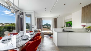 A modern kitchen with white cabinets, a dining area with red chairs, and a large window offering a city view. - Rausion Luxury Apartments (Dubrovnik)