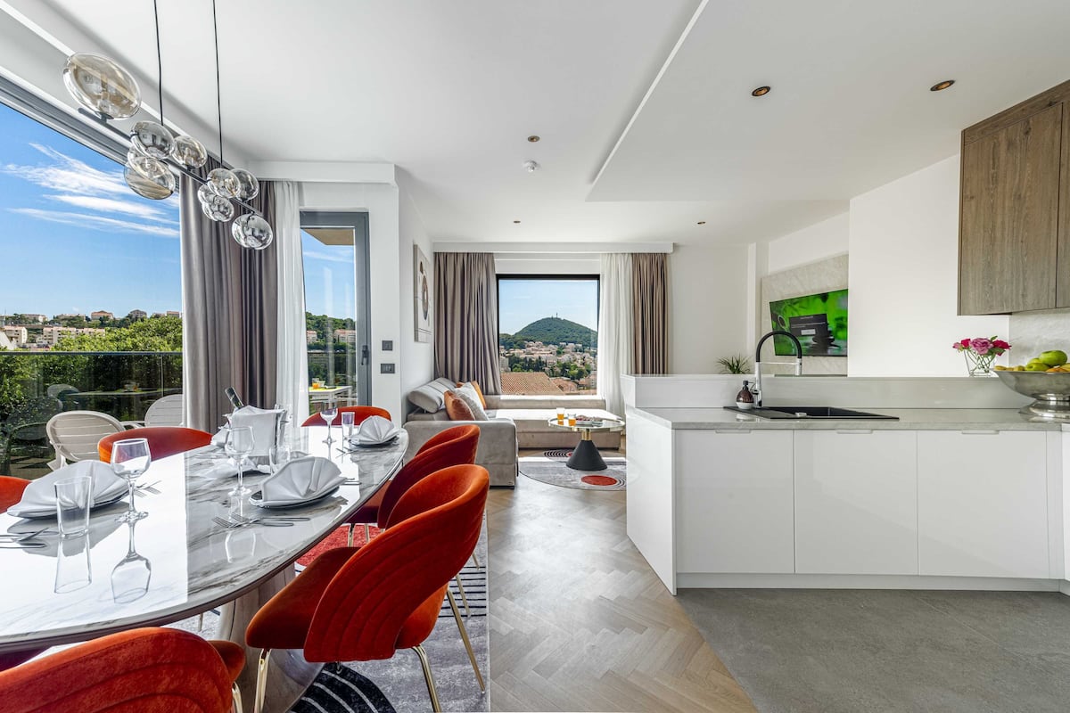 Une cuisine moderne avec des meubles blancs, un coin repas avec des chaises rouges et une grande fenêtre donnant sur la ville.