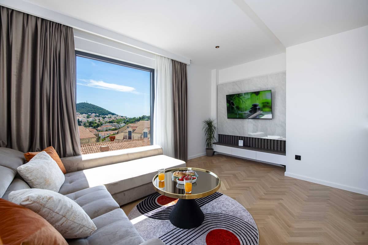 Un salon moderne avec une grande fenêtre, un canapé, une table basse avec un plateau de petit-déjeuner et un téléviseur fixé au mur.
