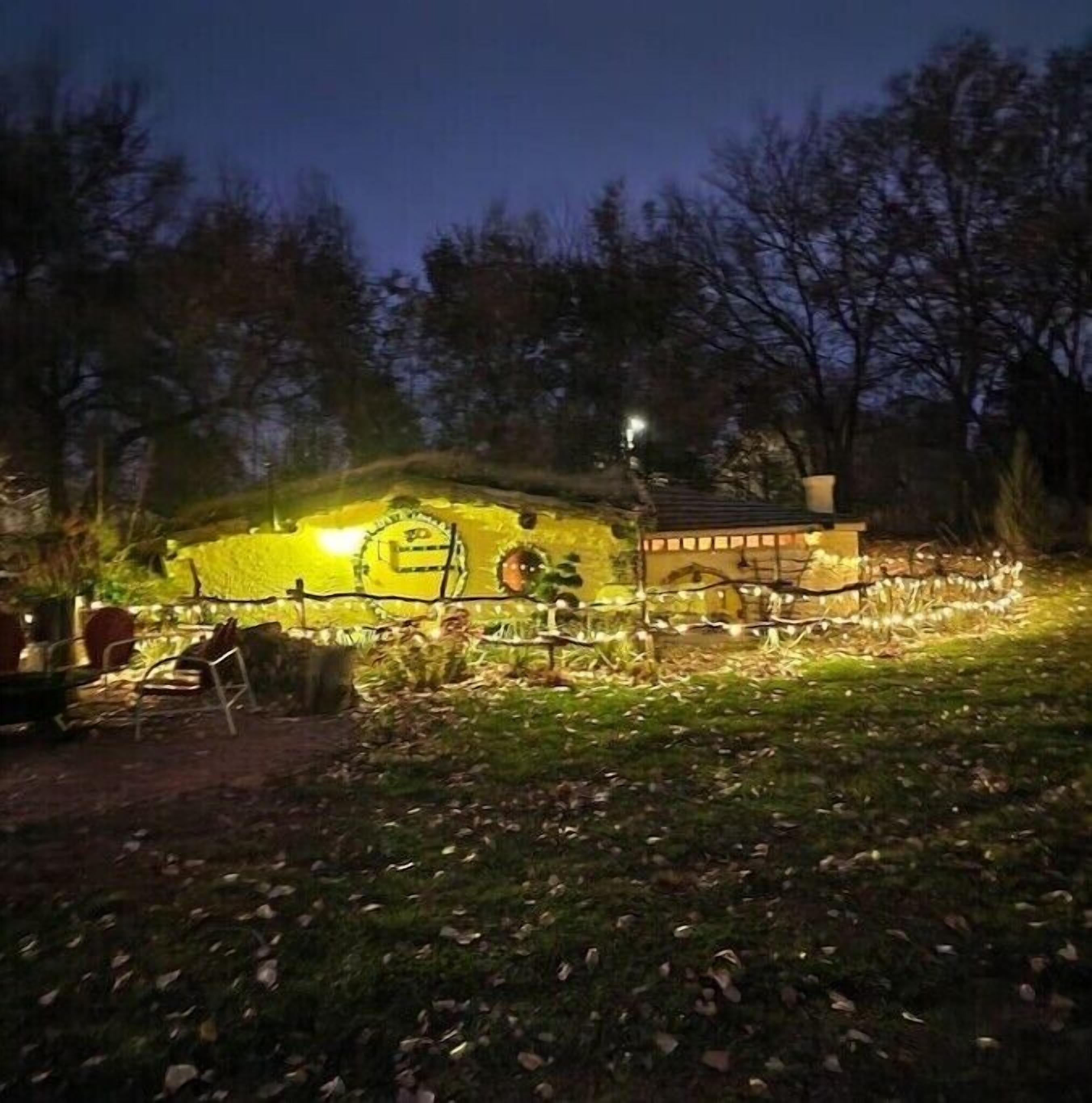 Una casa pequeña e iluminada con puerta y ventanas visibles, rodeada de una valla y árboles.