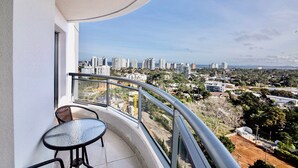 Un balcón con una mesa y sillas de vidrio, con vista a un paisaje urbano con edificios altos y áreas verdes.