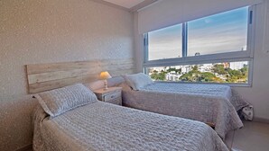 Un dormitorio con dos camas, una mesita de noche con lámpara y un ventanal con vistas a la ciudad.