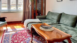 Un salon avec un canapé, une table basse en bois, un tapis à motifs, un radiateur et une fenêtre avec des rideaux.