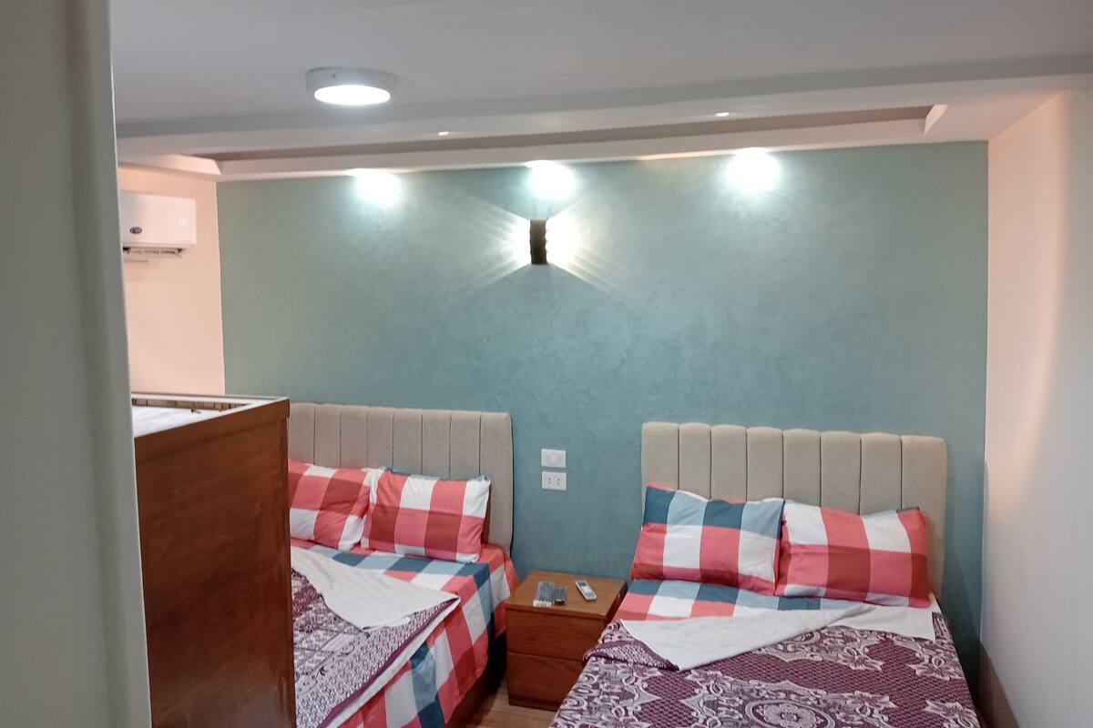 Ein Zimmer mit zwei Betten, jeweils mit kariertem Überwurf und Kopfteil. Ein Nachttisch mit Lampe und Telefon steht daneben. Die Wände sind blau und es gibt eine Deckenleuchte.