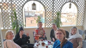Un grupo de personas sentadas alrededor de una mesa en un espacio exterior cubierto con estructura de celosía y vistas a edificios al fondo.