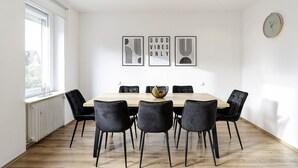 Une salle à manger avec une table en bois et six chaises noires. Trois œuvres d’art encadrées ornent le mur, et une horloge est placée à droite.