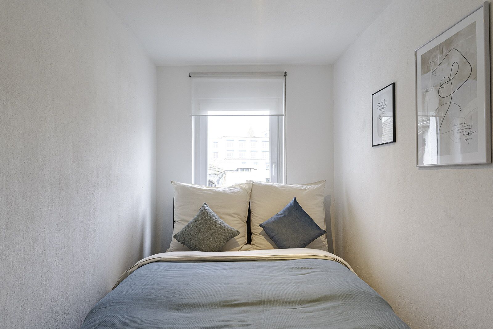 Ein Schlafzimmer mit einem Bett, zwei Kissen, einem Fenster und zwei gerahmten Kunstwerken an der Wand.