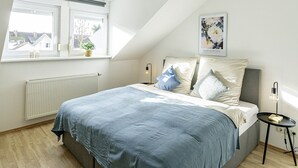 Une chambre avec un plafond en pente, un grand lit, une table de chevet, un radiateur et une fenêtre donnant sur des immeubles.