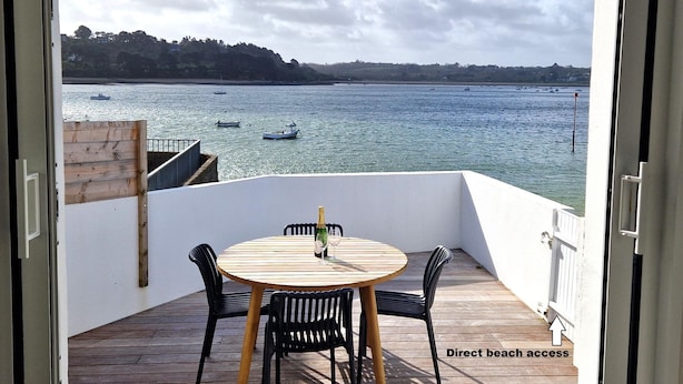 Een balkon met een houten tafel en stoelen, een champagnefles en een glas. Het balkon heeft uitzicht op een wateroppervlak met boten en een verre kustlijn.