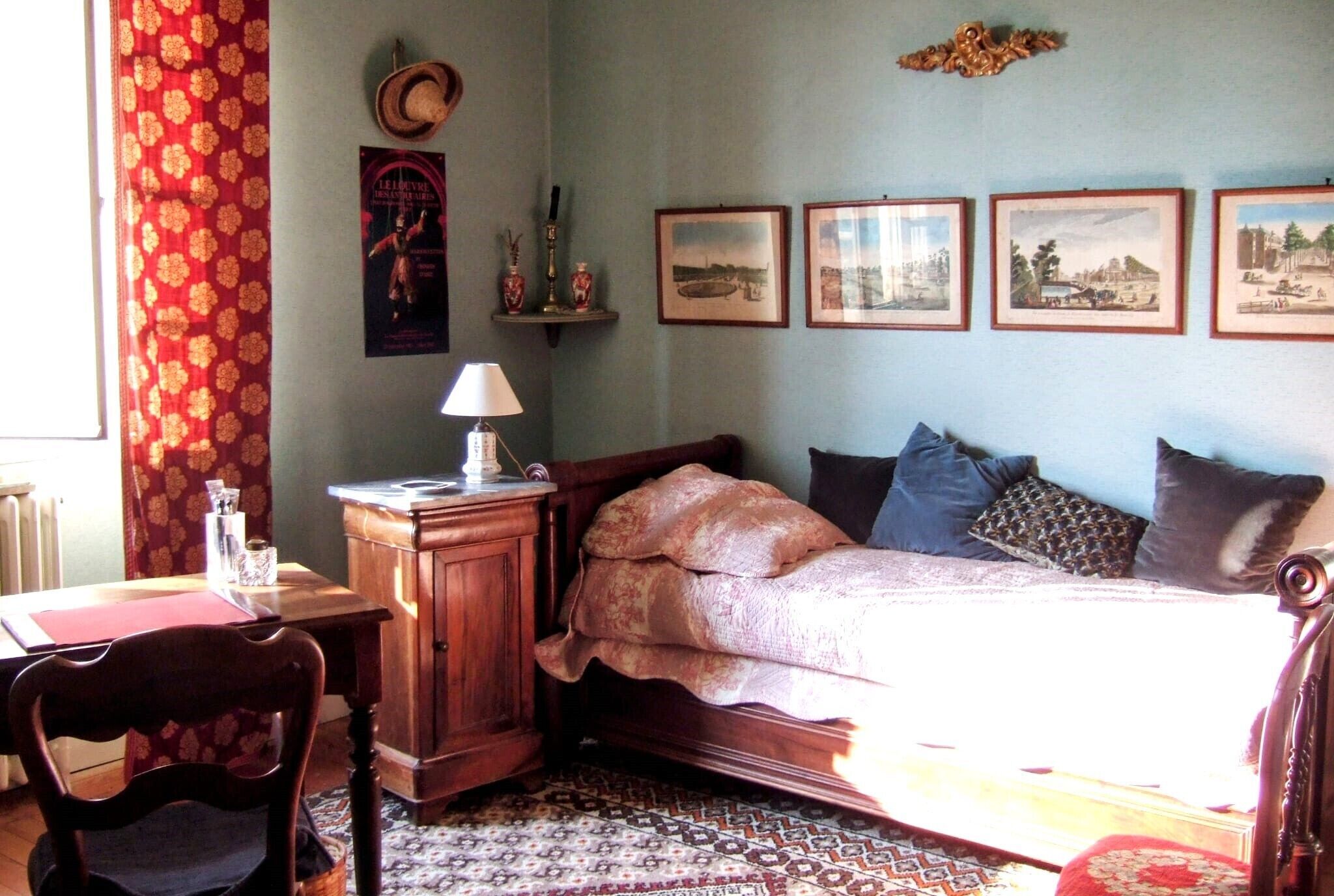 A bedroom with a wooden bed, a desk with a lamp, a chair, and a patterned rug. There are framed pictures on the wall and a window with curtains.