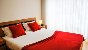 Un lit bien fait, avec une couverture rouge et des oreillers, une tête de lit en bois, une lampe de chevet et une fenêtre ornée de rideaux légers.