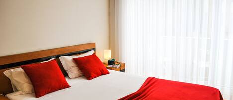 Una cama bien hecha con una colcha y almohadas rojas, cabecera de madera, lámpara de noche y una ventana con cortinas translúcidas.