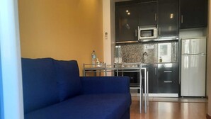 Un salon moderne avec un canapé bleu, une petite table à manger et un coin cuisine doté de meubles sombres et d’appareils électroménagers en acier inoxydable.