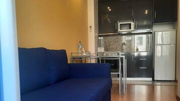 Un salon moderne avec un canapé bleu, une petite table à manger et un coin cuisine doté de meubles sombres et d’appareils électroménagers en acier inoxydable.