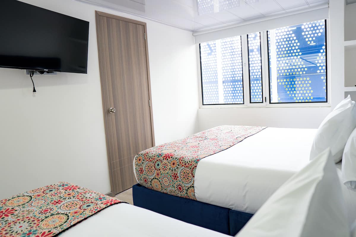 Une chambre d’hôtel avec un lit, un téléviseur fixé au mur, une fenêtre donnant sur des immeubles et une porte.