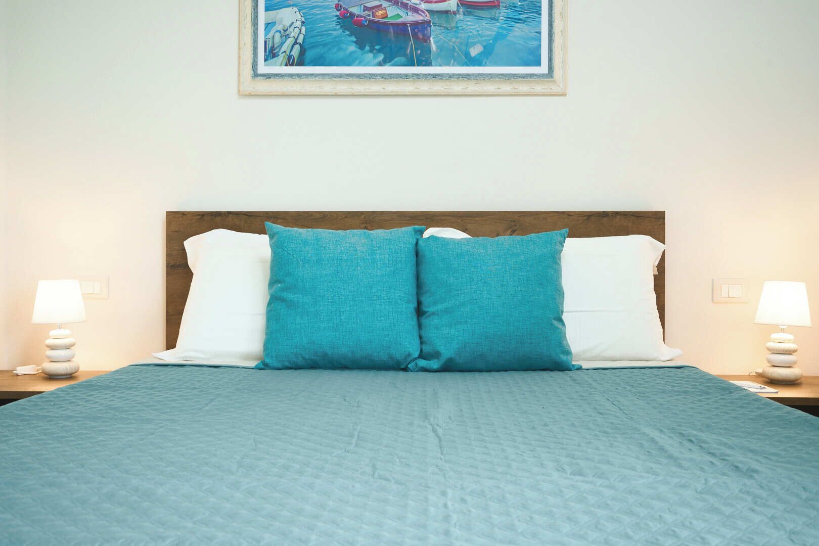 Un lit bien fait, recouvert d’une couvre-lit bleue, avec deux oreillers blancs et deux oreillers turquoise. Deux lampes de chevet sont placées de chaque côté du lit.