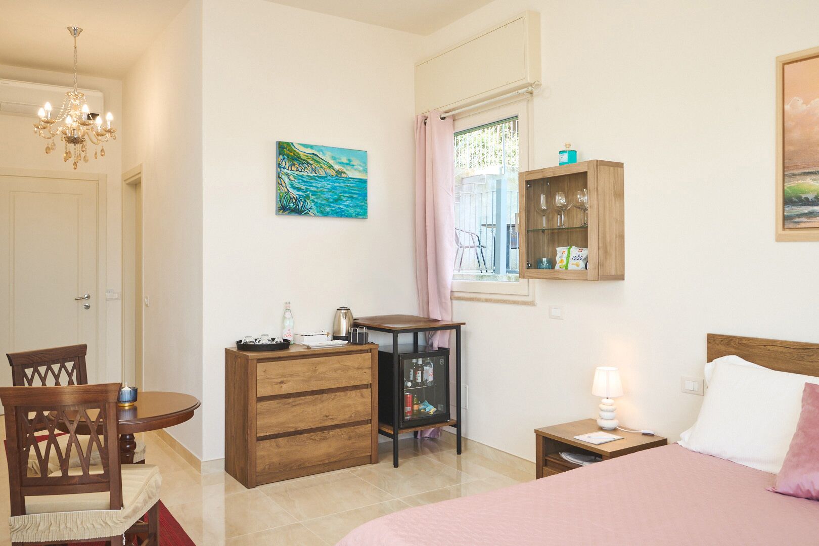 Une chambre à coucher comprenant un lit, un bureau avec une chaise, une commode, une table de chevet avec une lampe, une fenêtre avec des rideaux et des œuvres d’art accrochées aux murs.