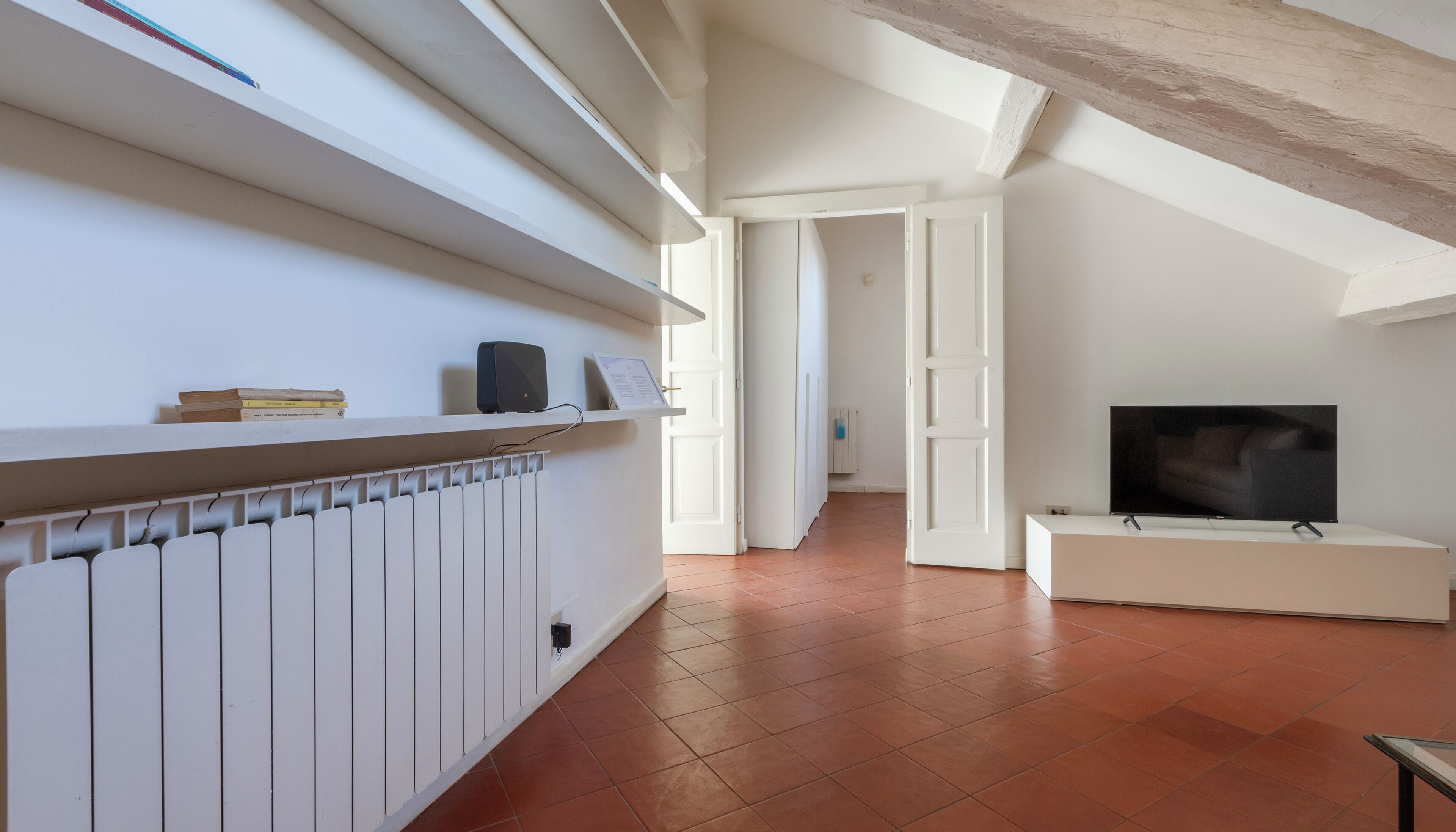 Uma sala de estar ampla com um radiador branco, uma TV de tela plana sobre um suporte branco e uma estante com livros e um alto-falante.