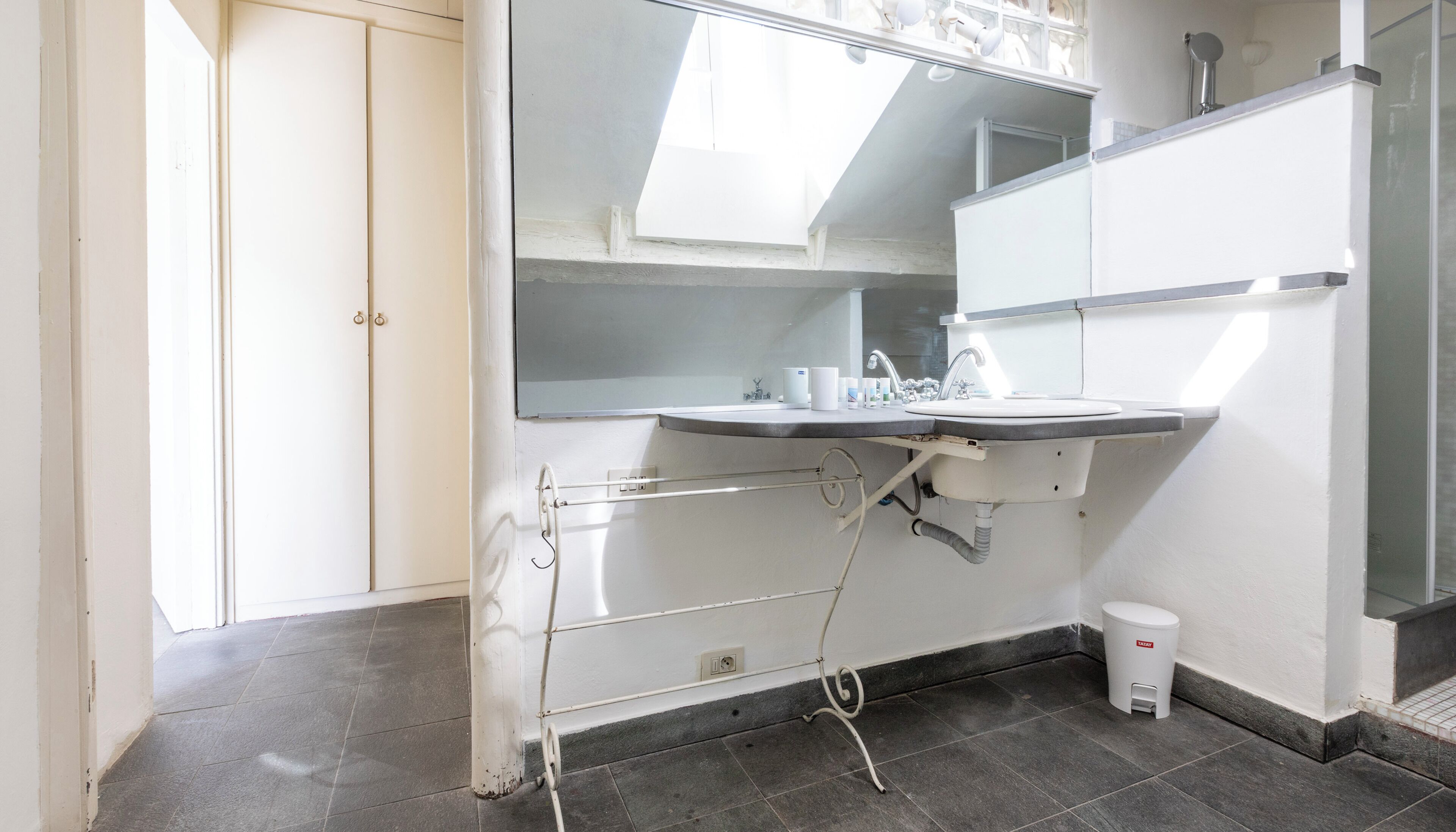 A modern bathroom with a white sink, mirror, and towel rack. There is a small trash bin and a shower area visible.