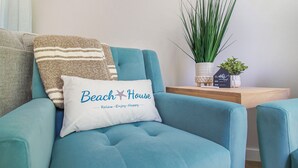 Un sillón color aguamarina con un cojín que dice "Beach House" y una pequeña mesita de madera con una planta y un letrero.