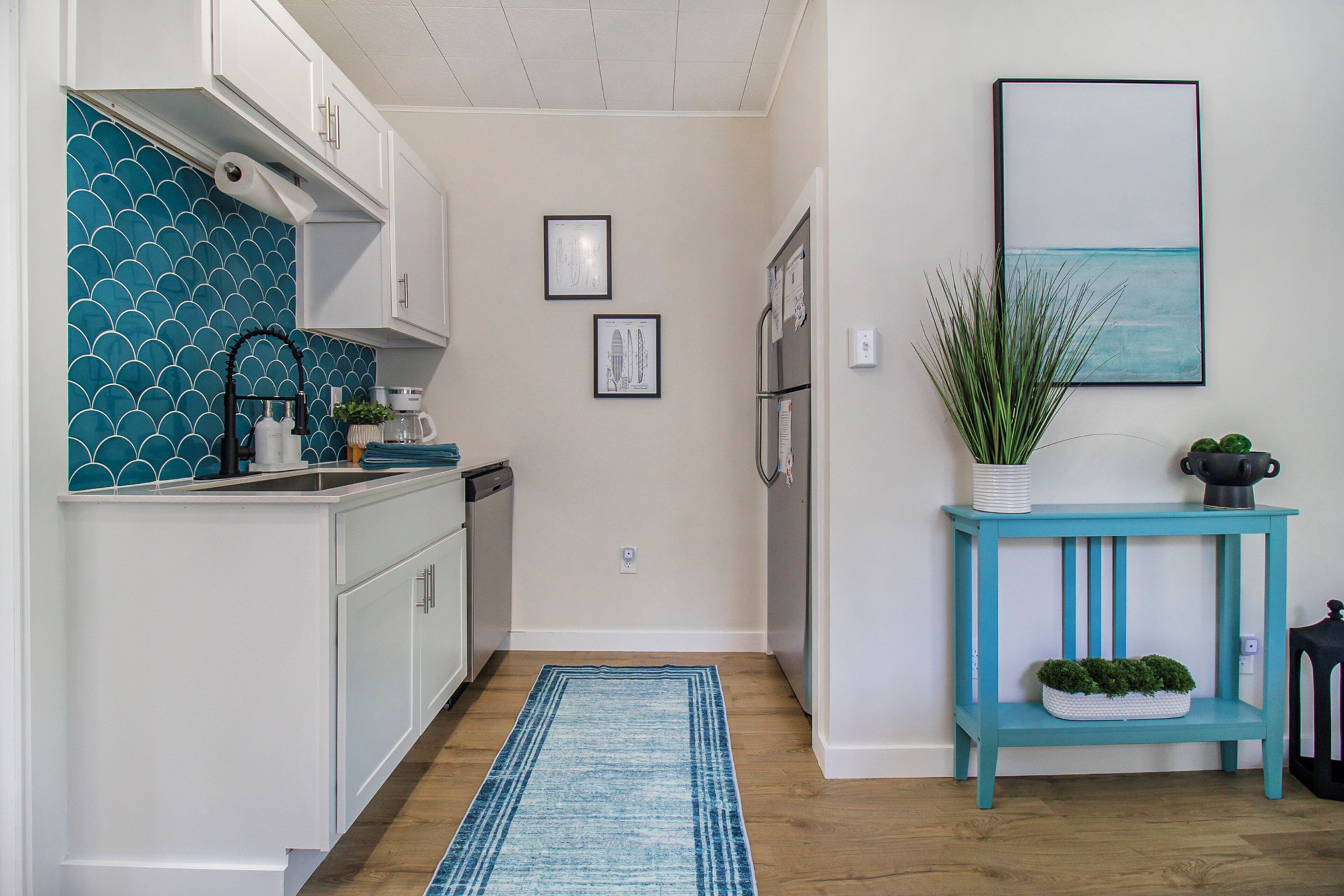 Une cuisine avec des meubles blancs, un pare-feu bleu et une console turquoise agrémentée d’une plante.