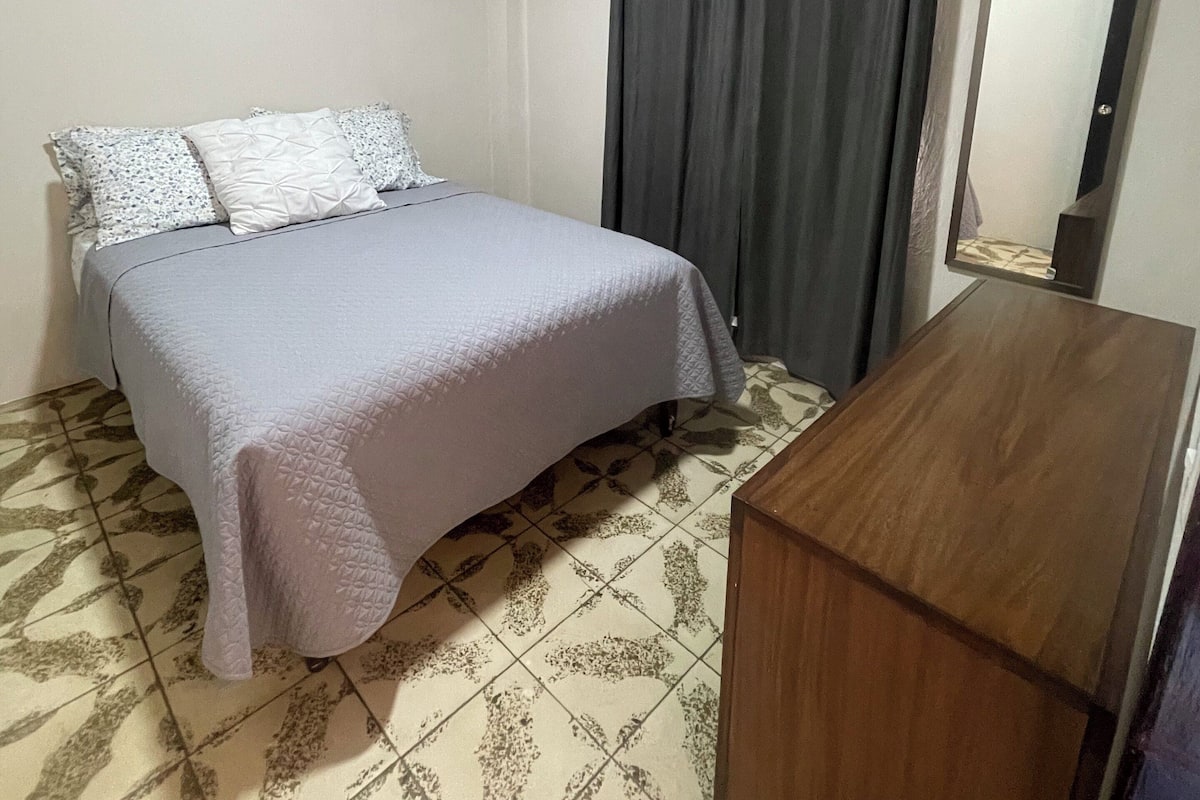 A bedroom with a bed, a wooden dresser, a mirror, and a window with curtains.