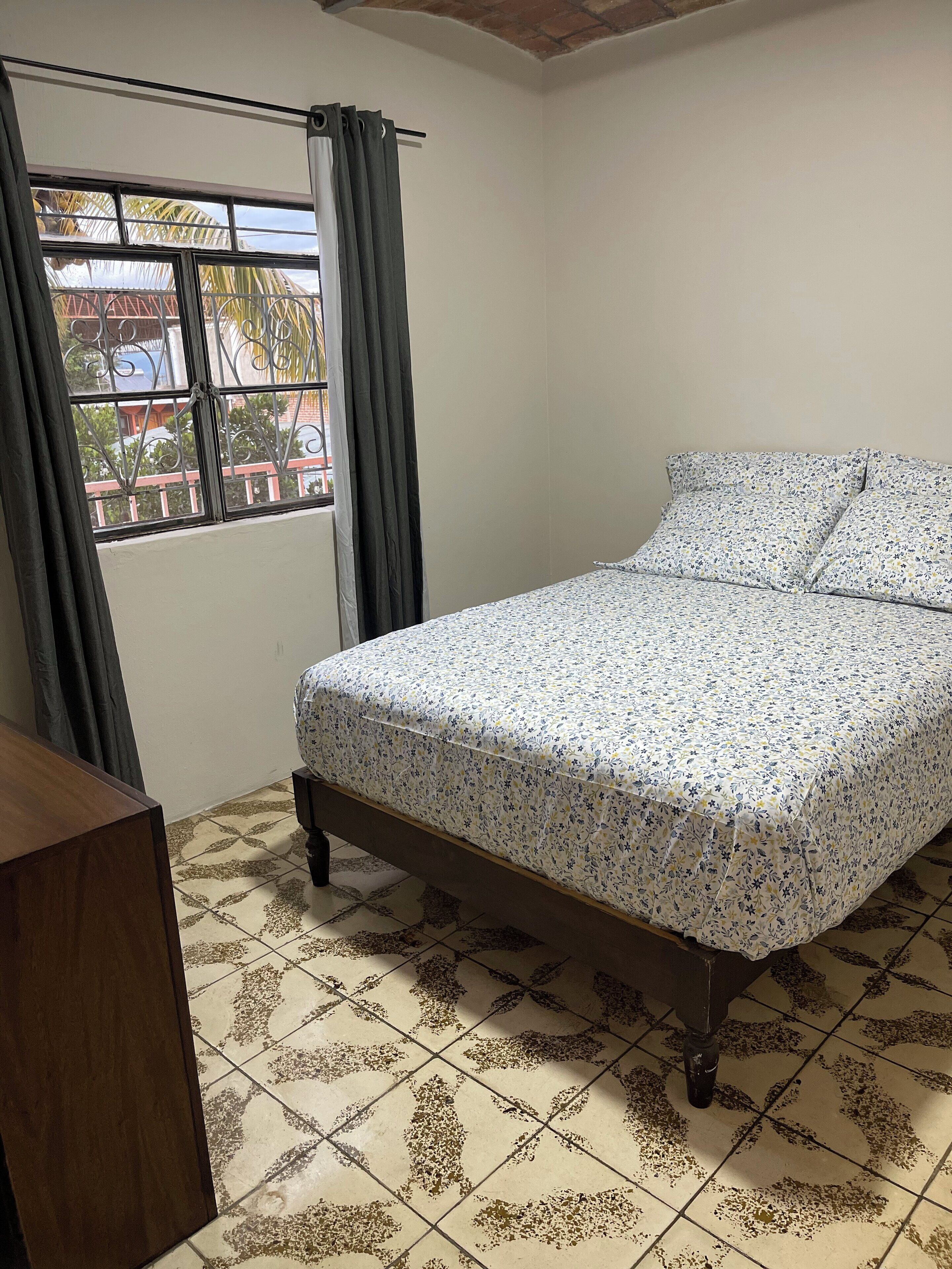A bedroom with a bed, curtains, a window, and a tiled floor.