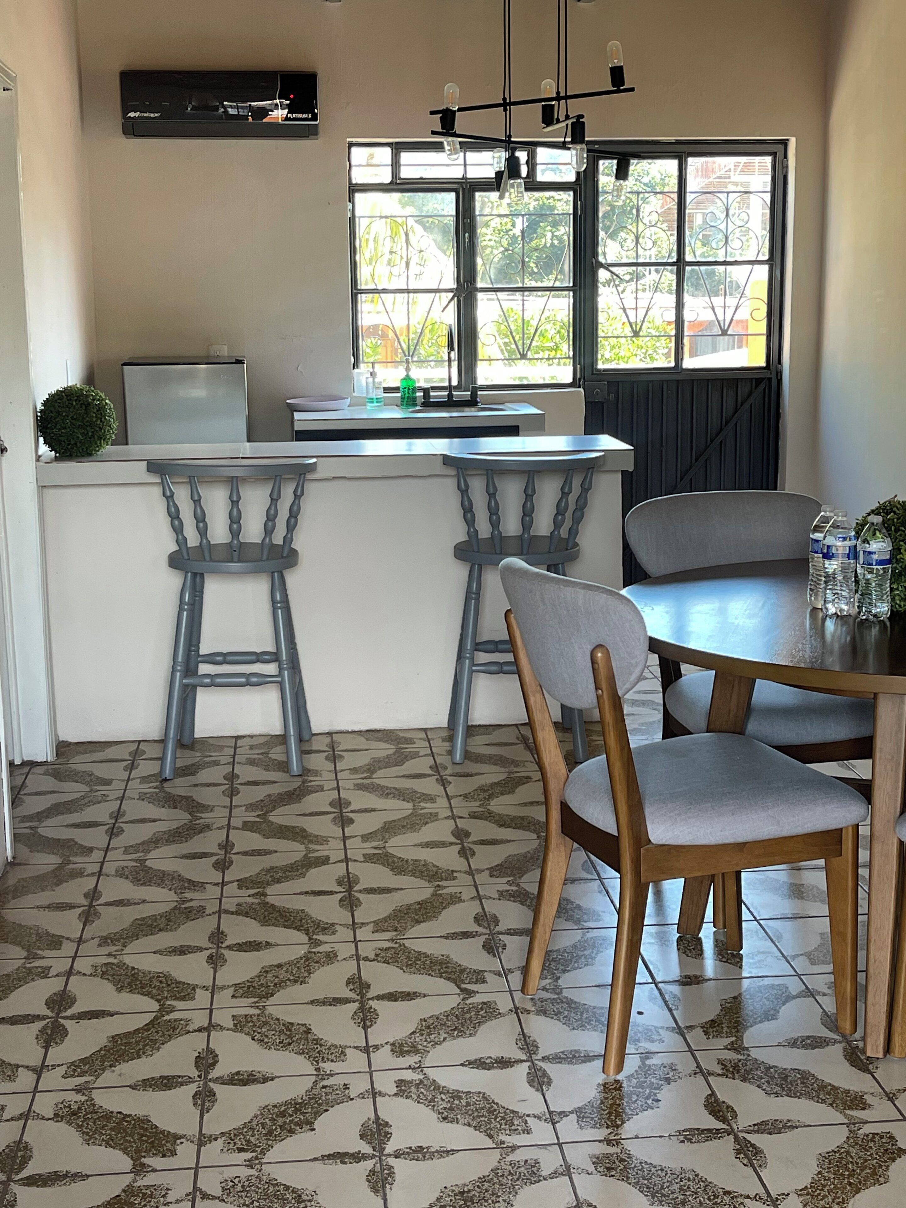 A compact kitchen with a refrigerator, a sink, and a window with decorative iron bars. There are two bar stools and a dining table with chairs.