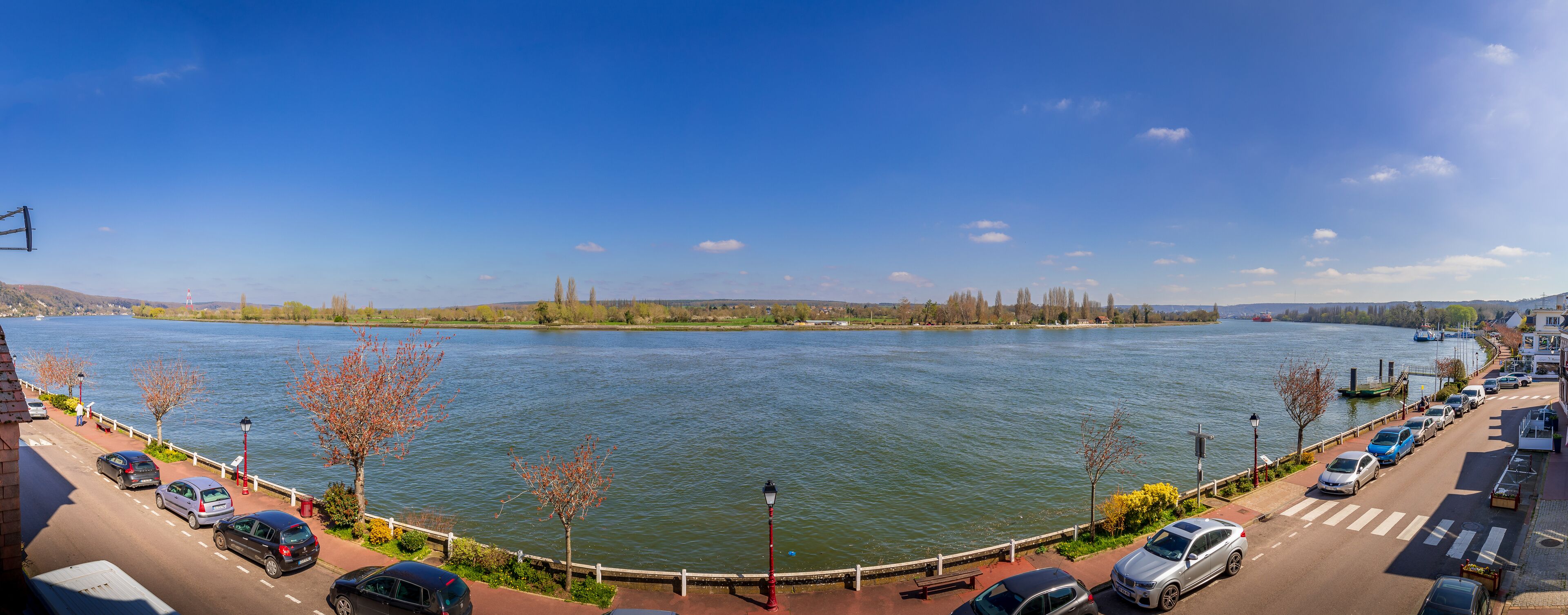 Blick auf einen Fluss mit Fußweg, parkenden Autos und blauem Himmel.