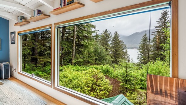 A room with large windows offering a view of a forest and lake. There are wooden shelves with books and a framed picture on the wall. - Serene Home with Ocean View   (Juneau)
