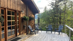 A wooden cabin with a deck, three Adirondack chairs, and a metal dog bowl. - Serene Home with Ocean View   (Juneau)