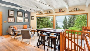 A modern living room with a large window, wooden ceiling beams, and a view of trees. - Serene Home with Ocean View   (Juneau)