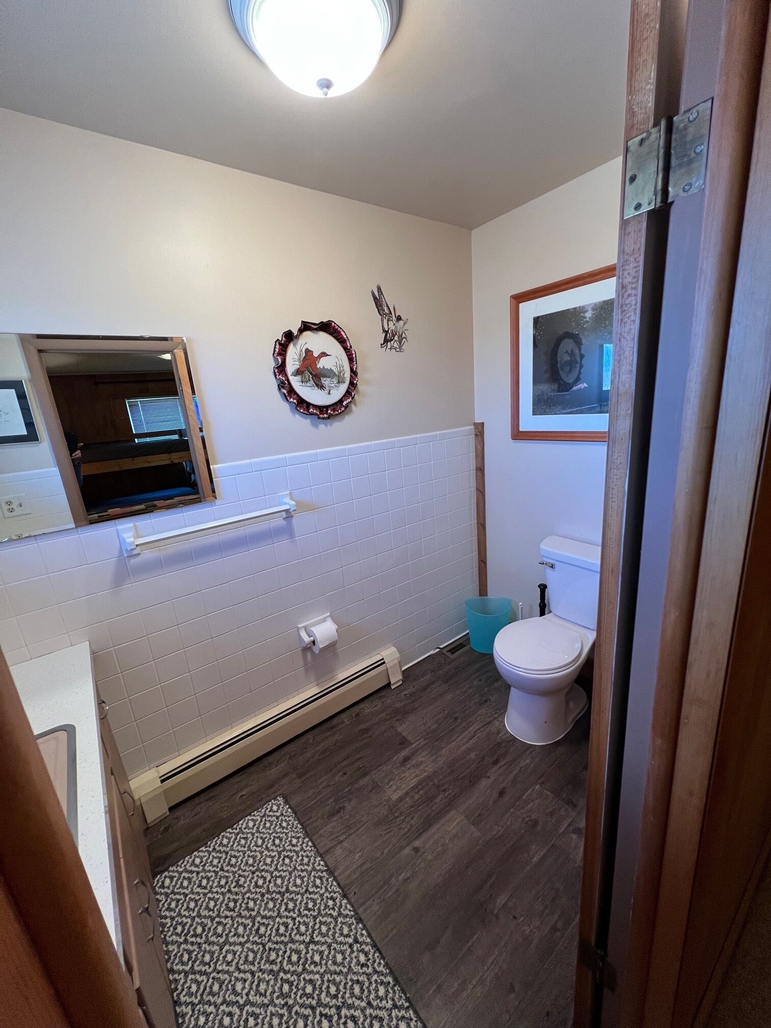 Un baño con pared de azulejos blancos, suelo de madera, inodoro, lavamanos, espejo y una pequeña ventana.