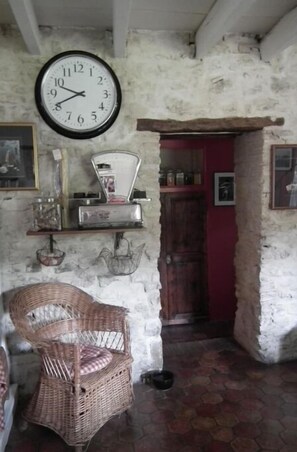 Une pièce rustique avec un fauteuil en osier, une horloge murale, une balance et une porte rouge.