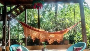 A hammock suspended between wooden beams, two turquoise chairs with cushions, and a small table with a vase.