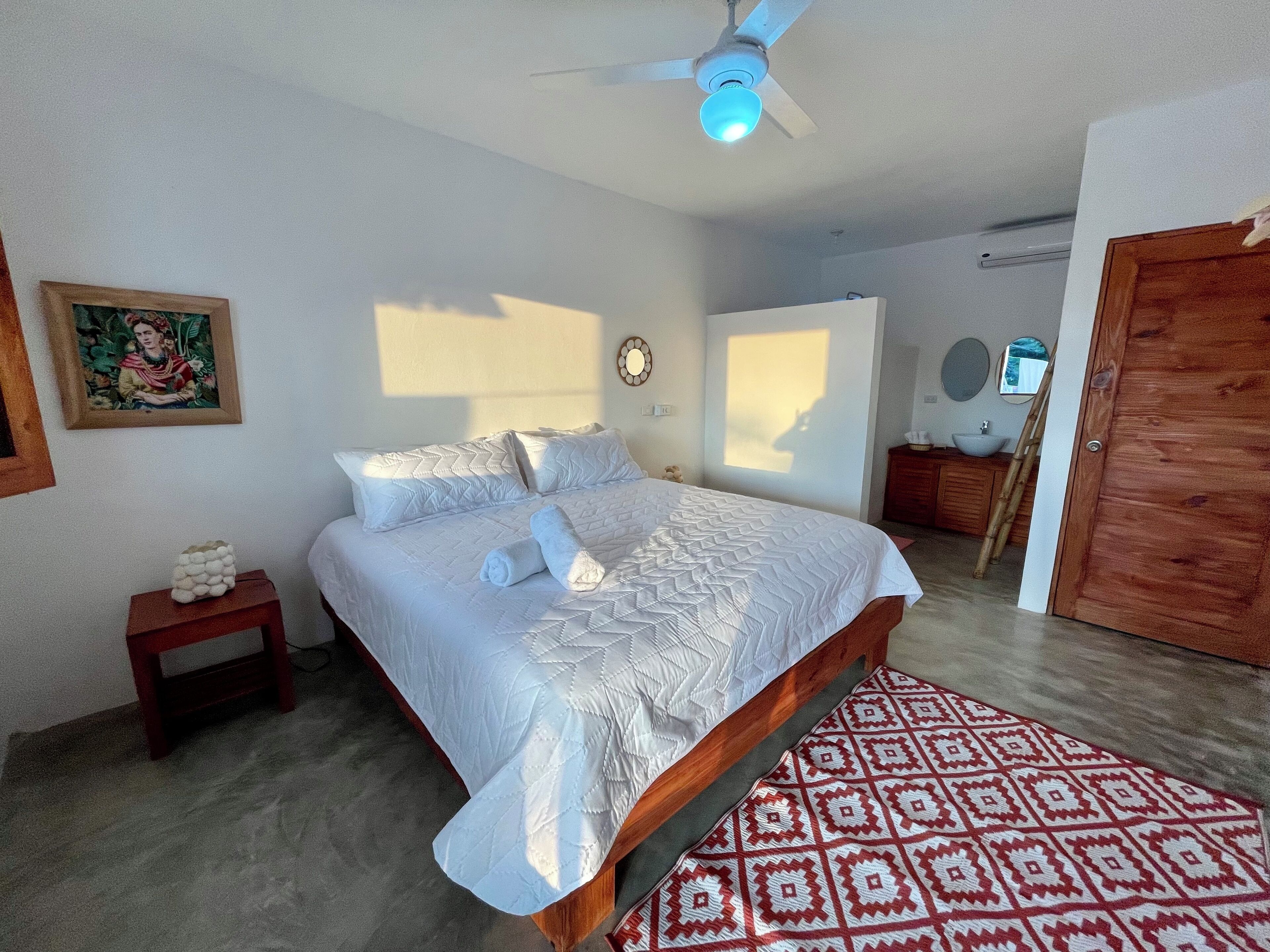 A bedroom with a bed, a bedside table, a nightstand, a wooden wardrobe, a mirror, and a patterned rug.