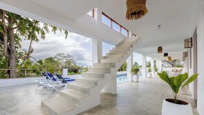 Àmplia zona interior amb una escala de formigó que condueix a un balcó, envoltada de plantes en testos i una piscina.