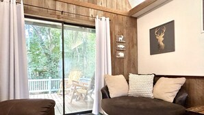 A cozy living room with a large window, a wooden deck, and a deer-themed wall decoration. - Doc's Cottage - A-Frame in Piggott, AR (Piggott)