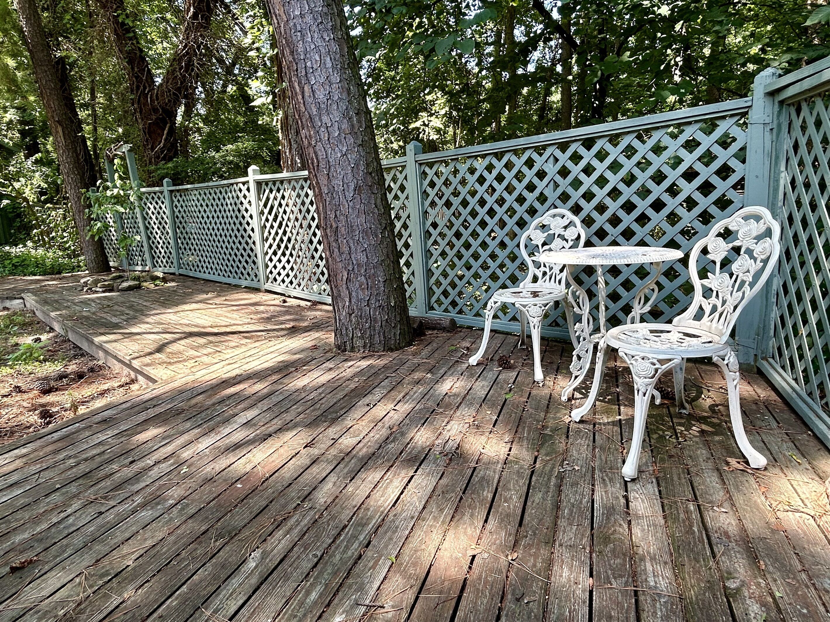 Una terraza de madera con dos sillas de metal blanco y una mesita pequeña, rodeada por un cerramiento de celosía y árboles.