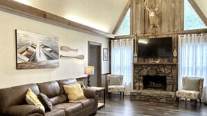 A cozy living room with a stone fireplace, leather sofa, and a mounted television. - Doc's Cottage - A-Frame in Piggott, AR (Piggott)
