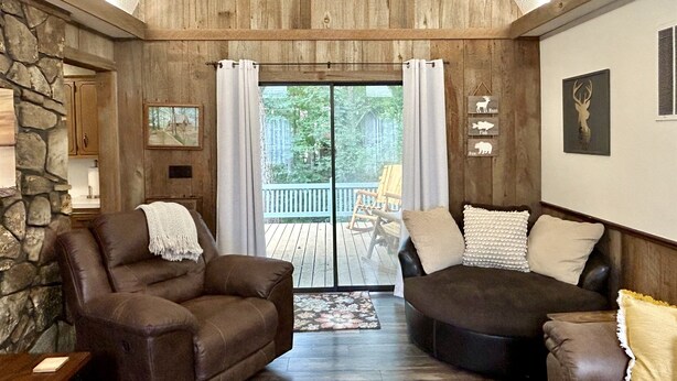 A cozy living room with two leather sofas, a stone wall, wooden ceiling, and a large sliding glass door leading to an outdoor deck. - Doc's Cottage - A-Frame in Piggott, AR (Piggott)