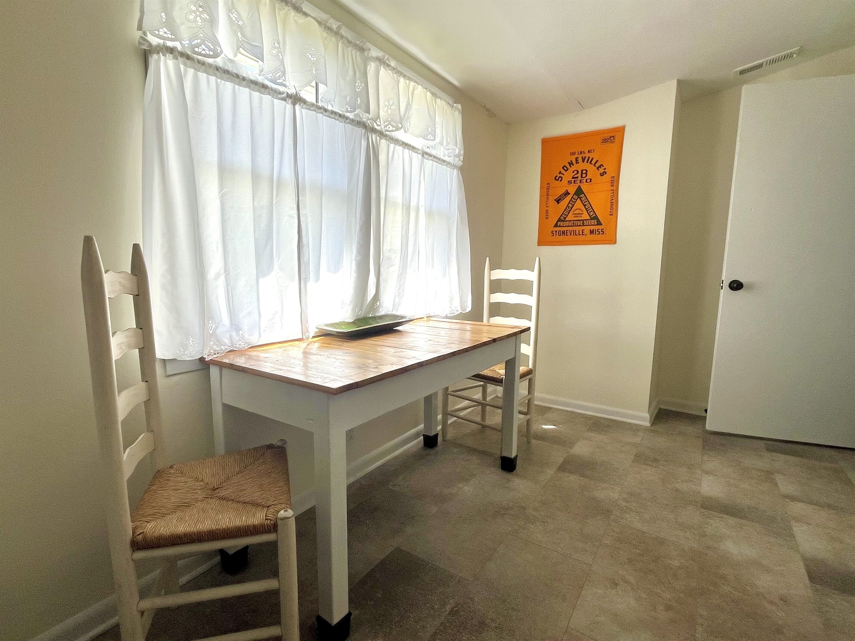 Un comedor con una mesa y dos sillas, una ventana con cortinas translúcidas y una puerta al fondo.