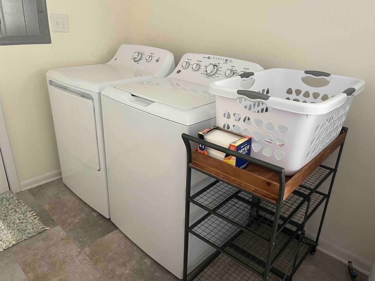 A laundry area with two white front-loading washing machines and a metal cart with a white laundry basket and detergent boxes.
