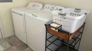 A laundry area with two white front-loading washing machines and a metal cart with a white laundry basket and detergent boxes.