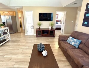Una sala de estar con un sofá marrón, una mesa de centro con piñas decorativas, un televisor montado en la pared y una zona de cocina con un refrigerador y armarios.